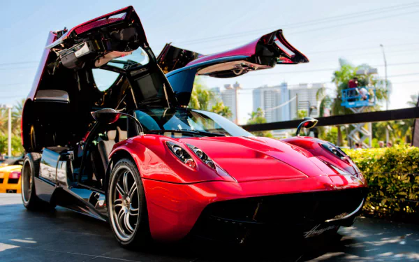 HD PC desktop wallpaper featuring a red Pagani Huayra with its gull-wing doors open, parked outdoors against a cityscape backdrop.