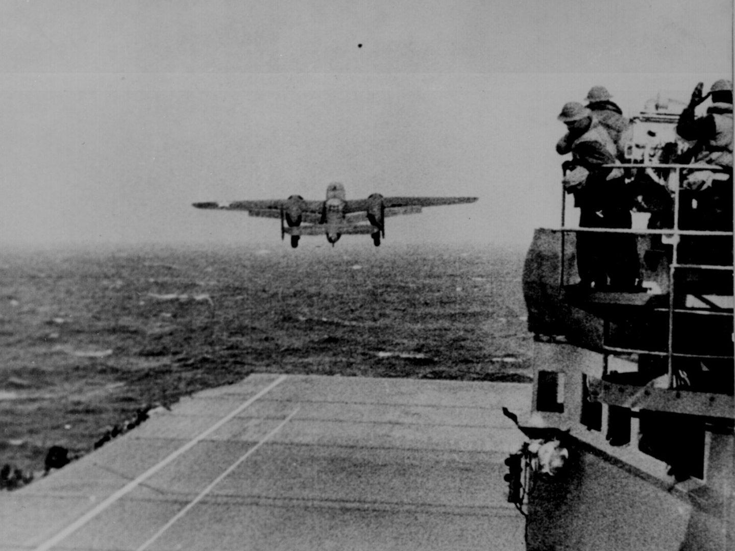 Black and white HD desktop wallpaper showing a North American B-25 Mitchell aircraft approaching an aircraft carrier deck with military personnel observing.