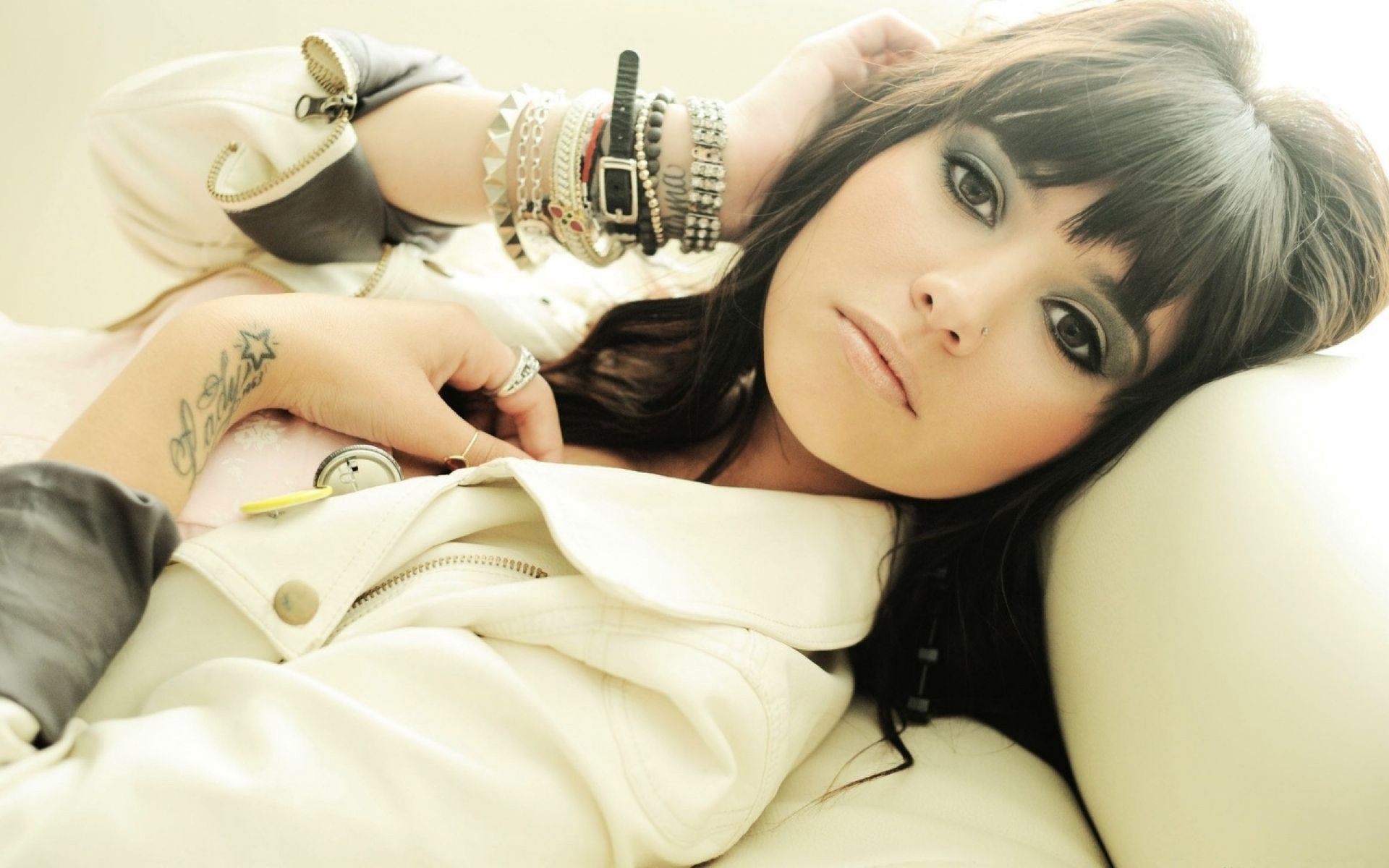 A stylish woman with dark hair and bold makeup reclines against a soft surface, adorned with bracelets. This HD image makes an eye-catching desktop wallpaper and background.
