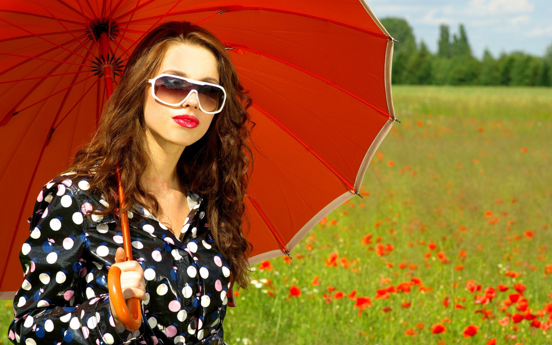 HD photography desktop wallpaper featuring a woman in a black polka dot dress holding a bright red umbrella in a vibrant green field with scattered red flowers.