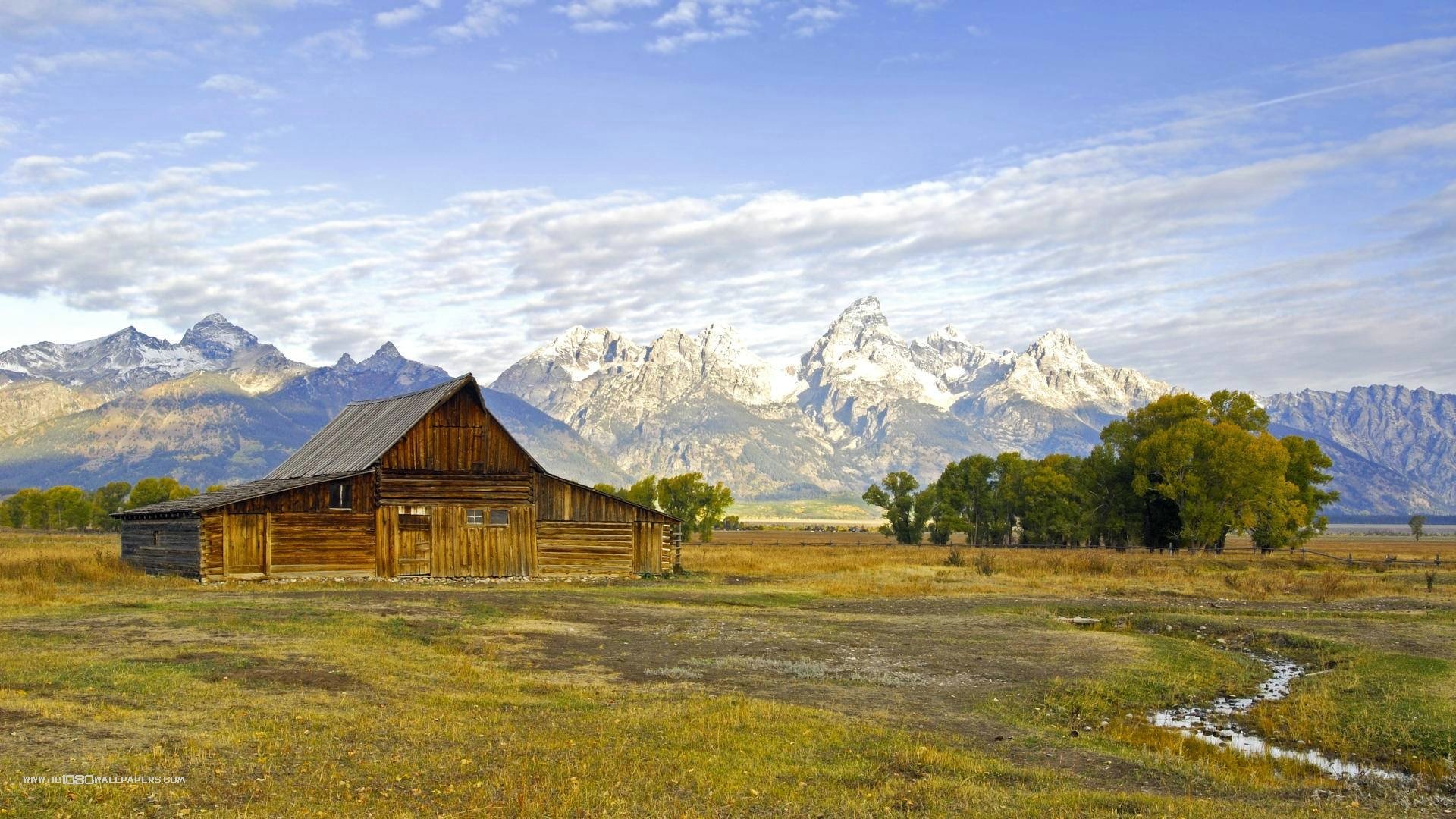 HD desktop wallpaper of a man-made wooden cabin set in a vast field with a backdrop of snow-capped mountains under a partly cloudy sky.