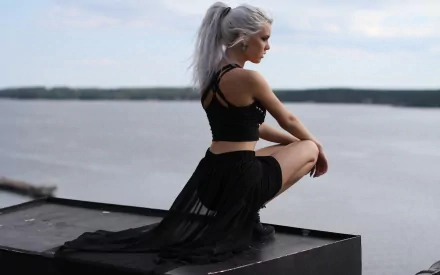 A woman with white hair styled in a ponytail, wearing earrings and a black outfit, sits contemplatively by a vast water body, suitable as an HD desktop wallpaper.