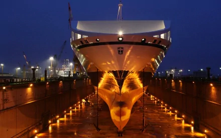 HD desktop wallpaper of a large ship in a man-made shipyard, illuminated at night with reflections on the dry dock floor under deep blue sky.
