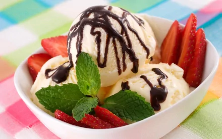 HD desktop wallpaper featuring a bowl of vanilla ice cream with chocolate syrup, fresh strawberries, and mint leaves on a colorful checkered cloth.