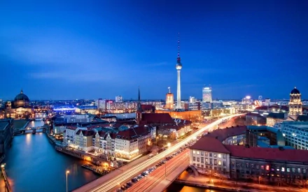 A stunning HD desktop wallpaper of Berlin at dusk, showcasing the city's skyline with illuminated buildings, the Berlin TV Tower, and a river reflecting glowing lights.