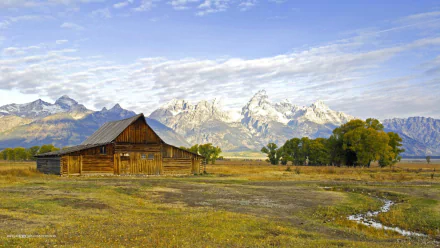 HD desktop wallpaper of a man-made wooden cabin set in a vast field with a backdrop of snow-capped mountains under a partly cloudy sky.