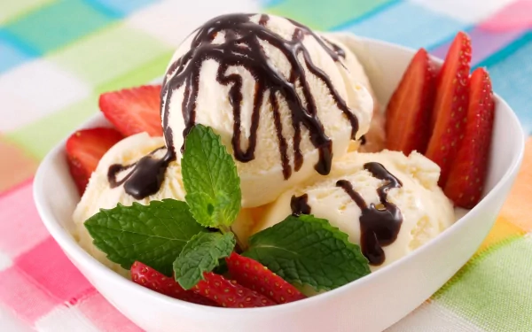 HD desktop wallpaper featuring a bowl of vanilla ice cream with chocolate syrup, fresh strawberries, and mint leaves on a colorful checkered cloth.