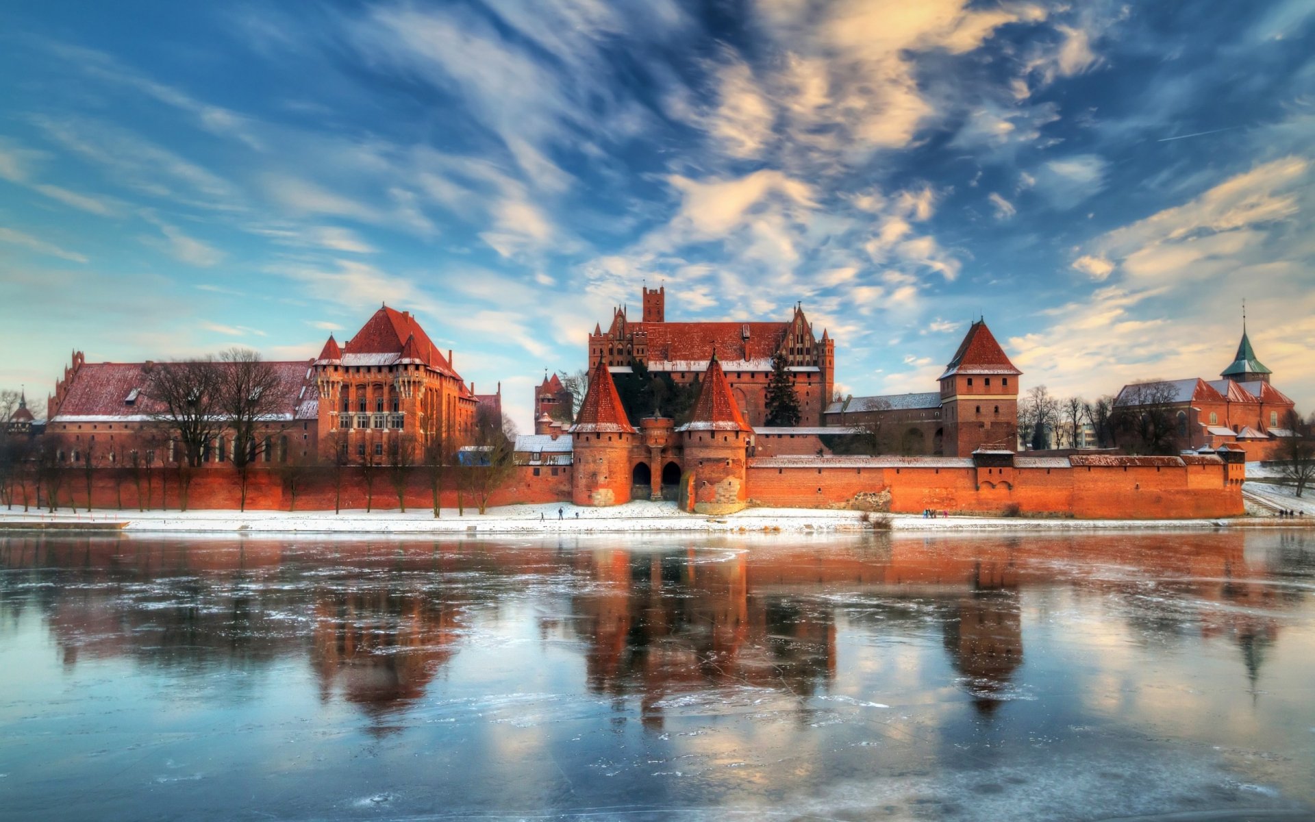 A stunning view of Malbork Castle, reflecting in the water beneath a vibrant sky, showcases its impressive medieval architecture, making for a captivating HD desktop wallpaper.