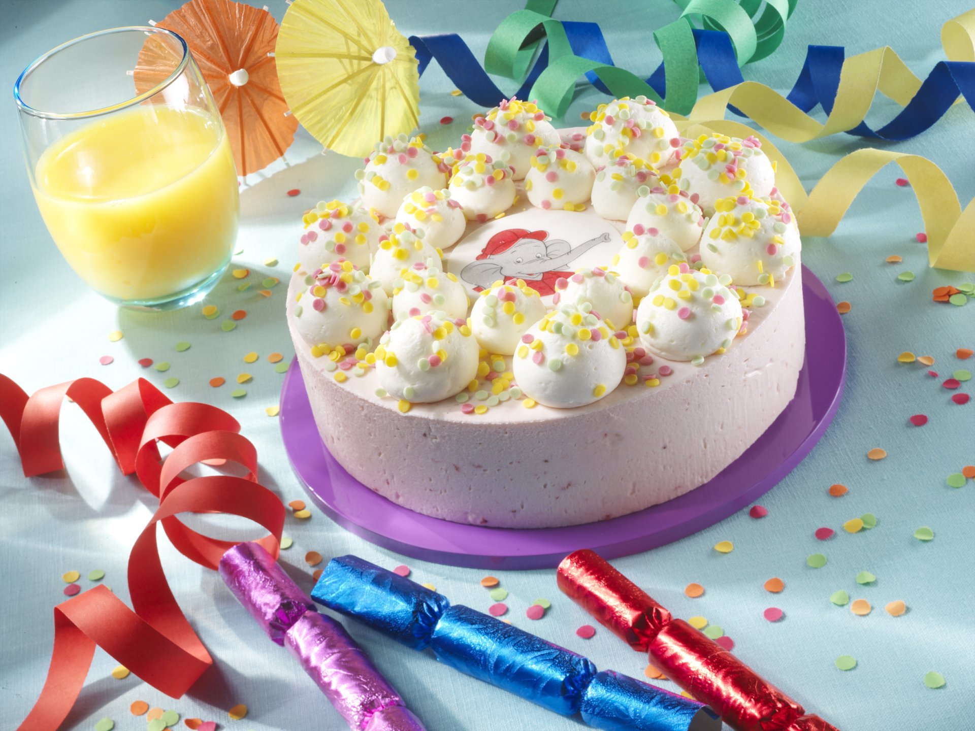 A colorful cake topped with creamy frosting and sprinkles, surrounded by festive decorations and a glass of juice, featured as a vibrant HD desktop wallpaper.