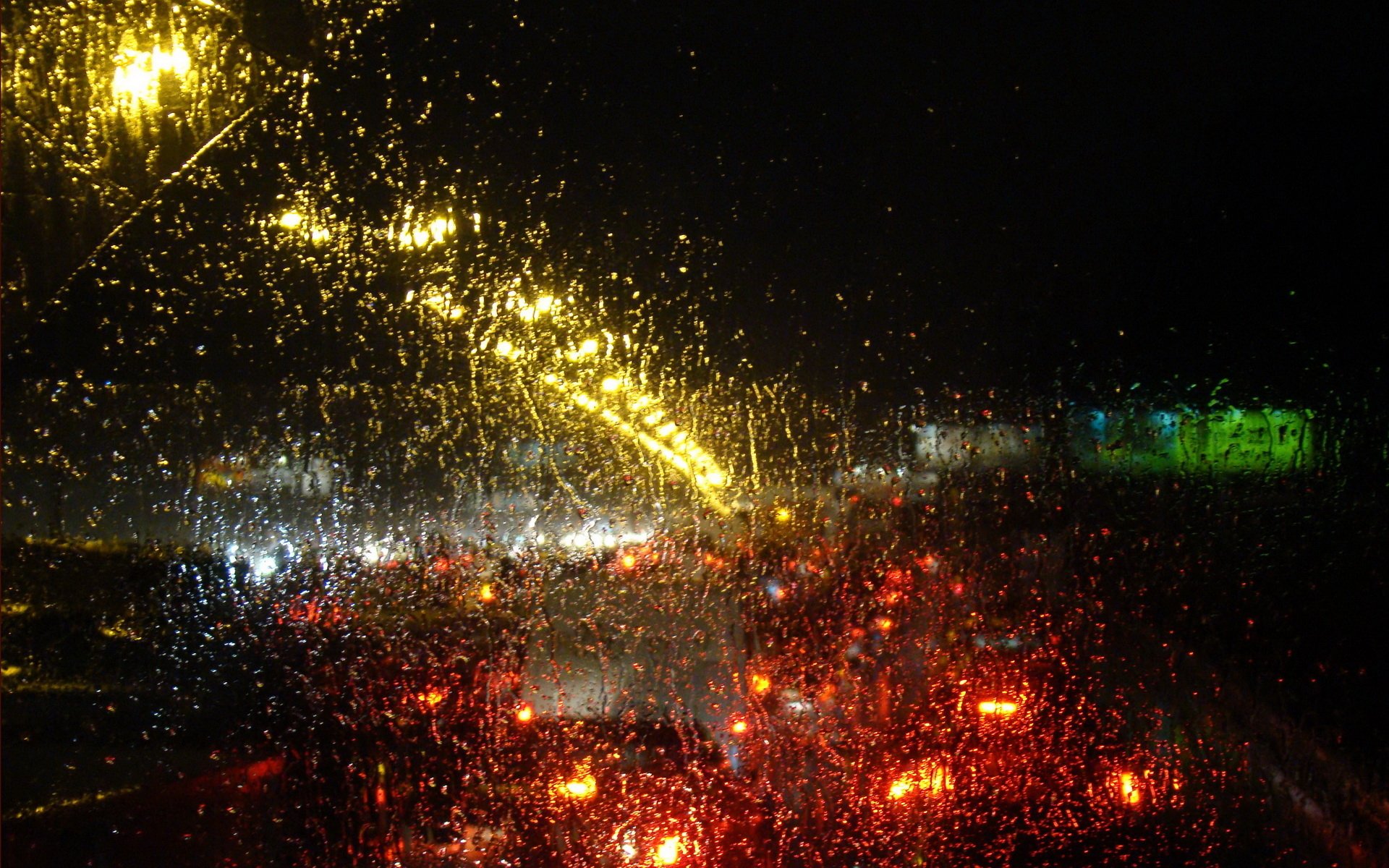 HD photography desktop wallpaper capturing raindrops on a car window with blurred red and yellow city lights in the background, creating a vibrant night scene.