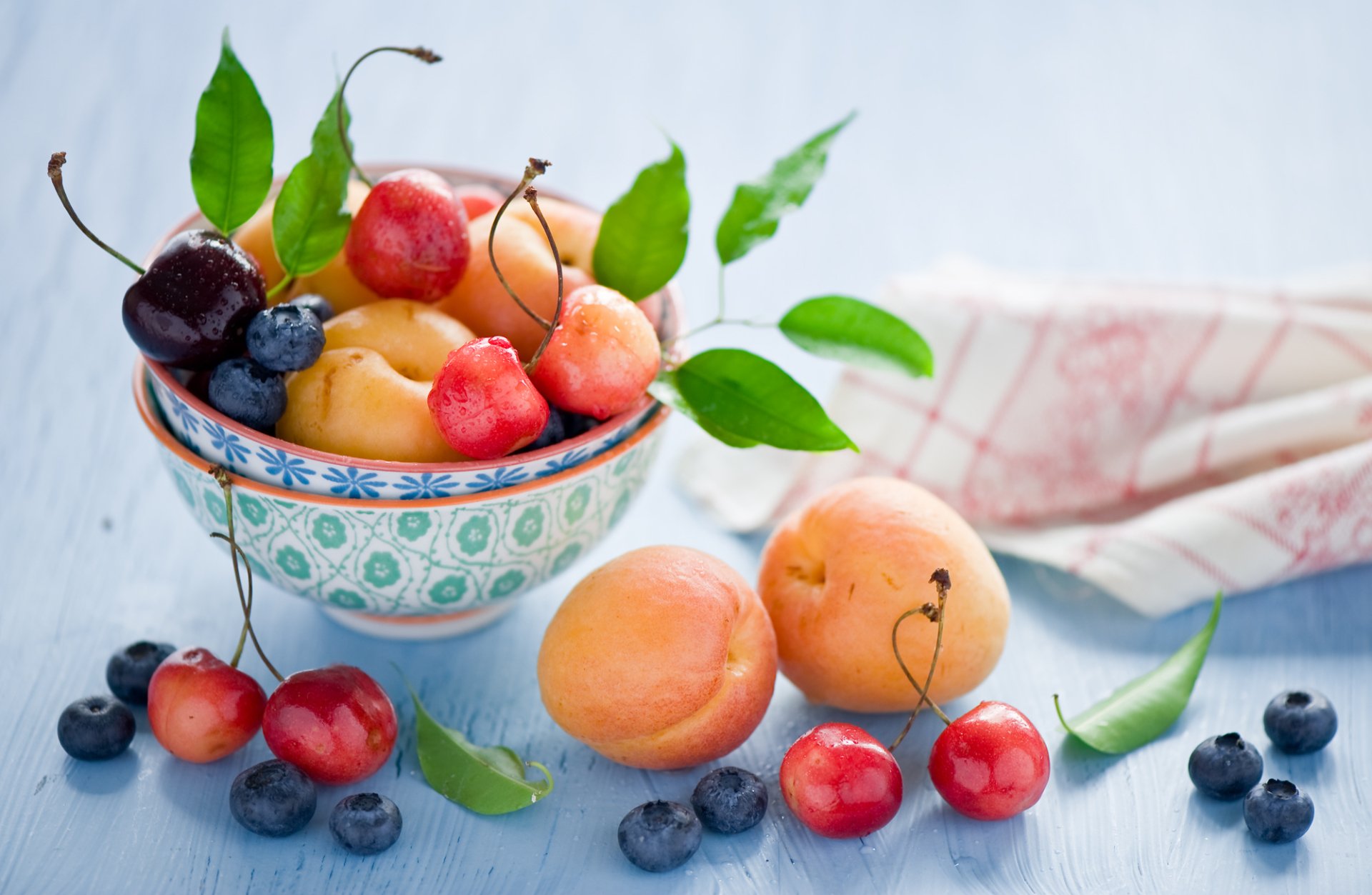 A colorful arrangement of fresh fruits, including cherries, blueberries, and apricots, displayed in a decorative bowl, creating a vibrant desktop wallpaper background.