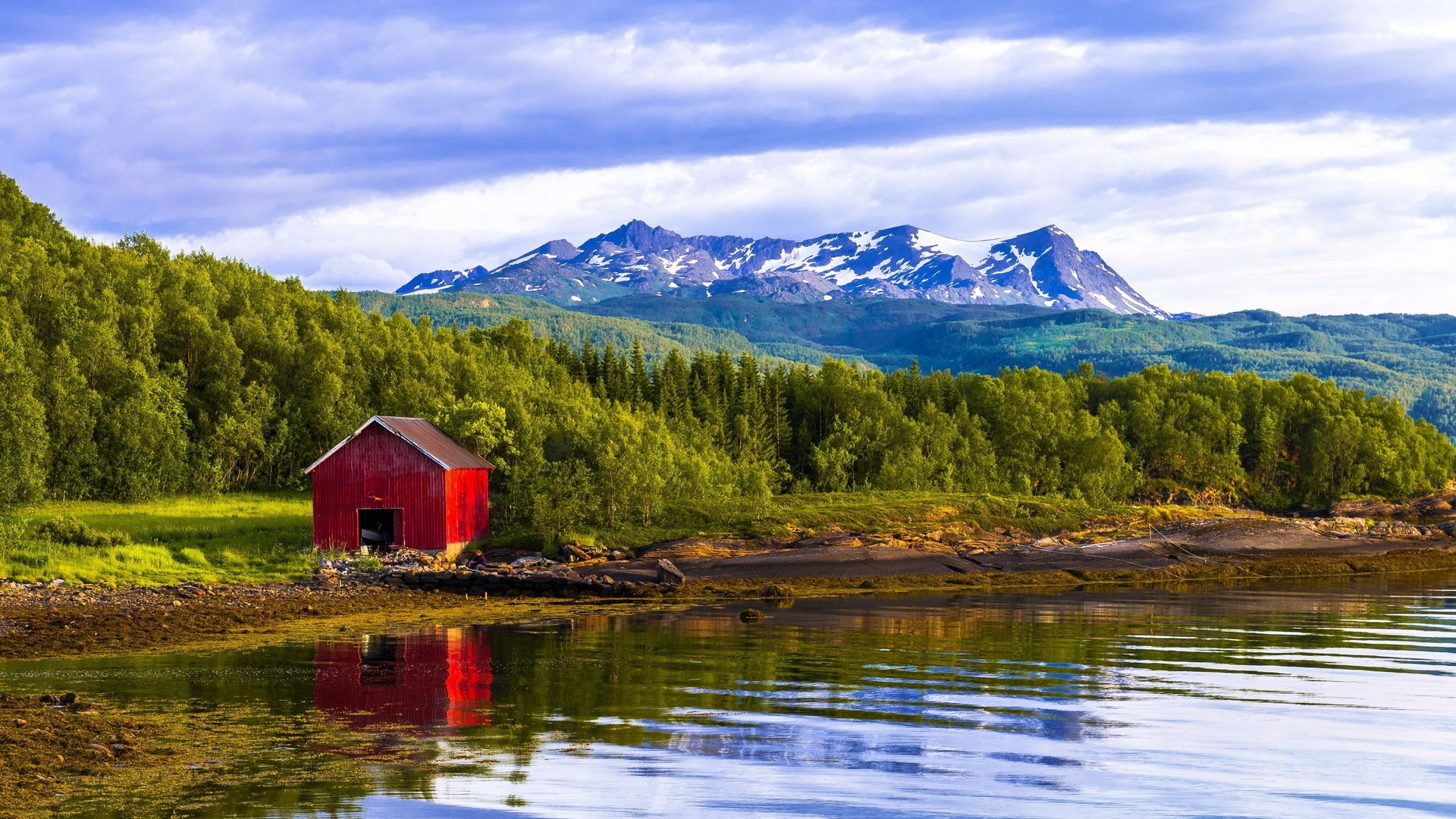 HD desktop wallpaper featuring a man-made red cabin by a calm lake, surrounded by lush green trees and distant snow-capped mountains under a cloudy sky.