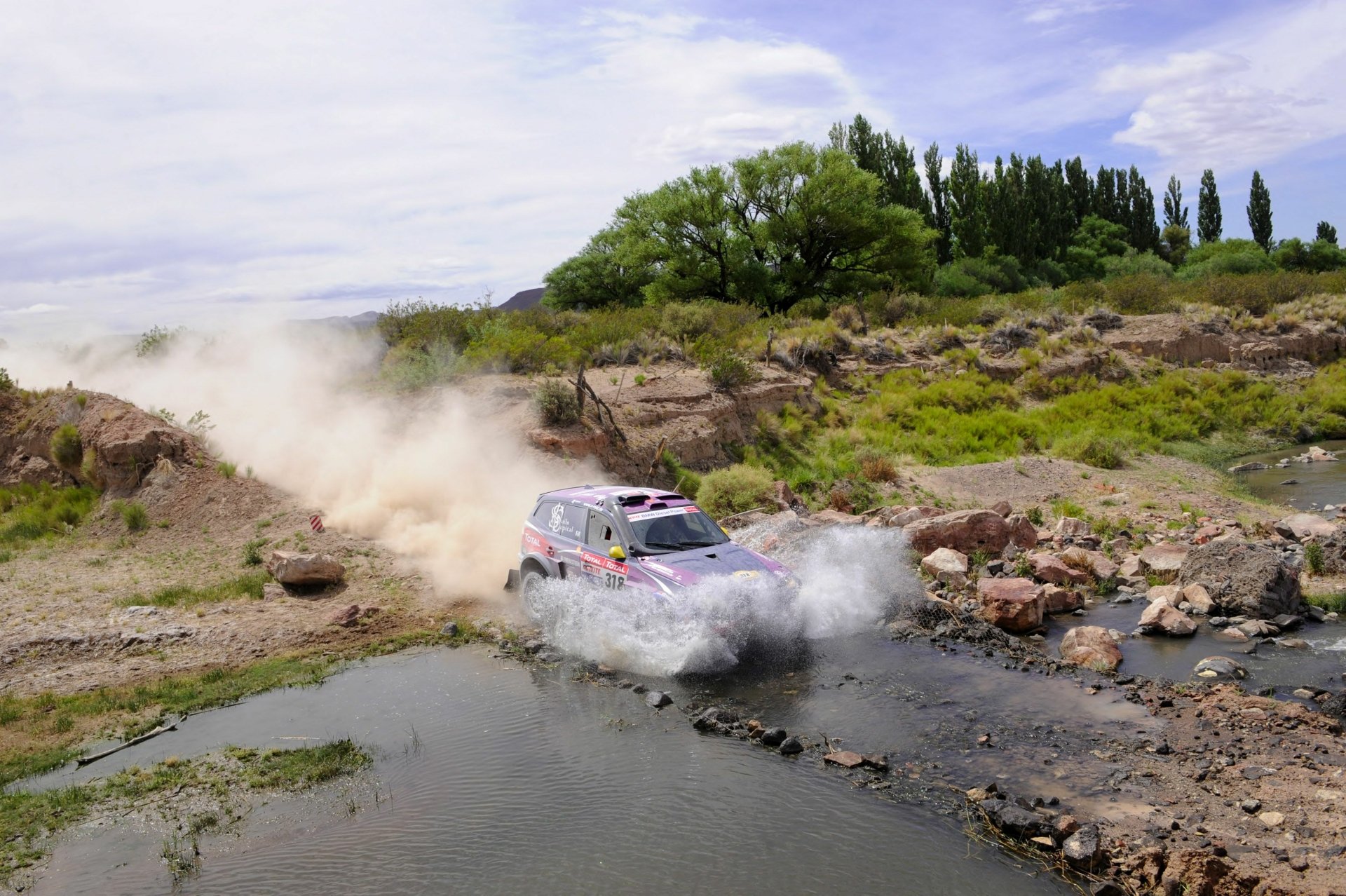 Rally BMW charging through a rocky stream, throwing spray and dust across rugged terrain — 2K Quad HD PC desktop wallpaper and background.