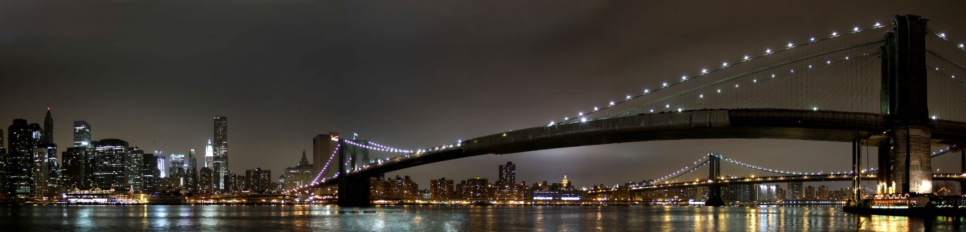 Night view of the illuminated Brooklyn Bridge spanning over water with the Manhattan skyline in the background, captured in 4K Ultra HD quality.