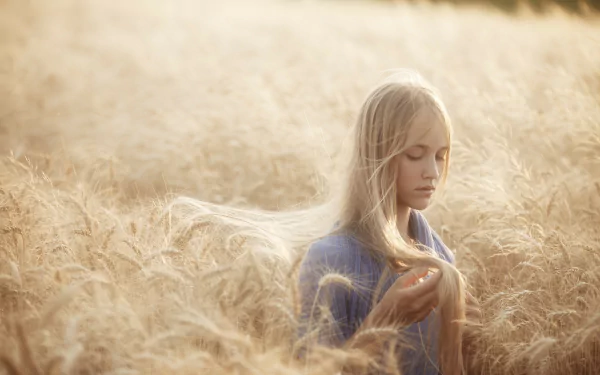 A serene woman stands in a golden field, gently holding her long hair as sunlight bathes the scene, creating a beautiful HD desktop wallpaper.