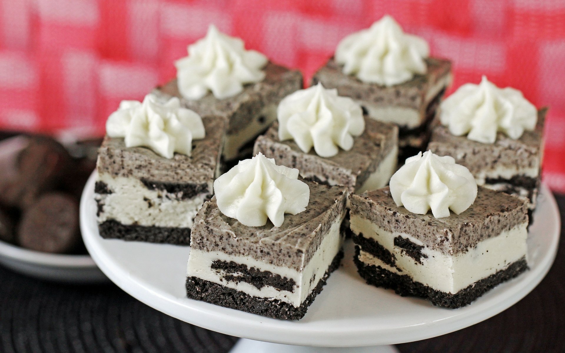 HD PC desktop wallpaper featuring delicious Oreo cream cake squares topped with whipped cream on a white serving plate against a red checkered background.