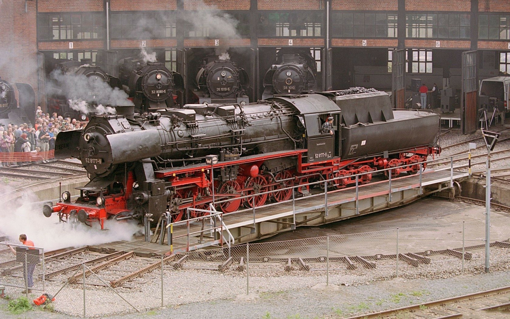A majestic steam train rotates on a turntable, surrounded by classic locomotives and an intrigued crowd, creating a captivating scene for a train-themed HD desktop wallpaper.