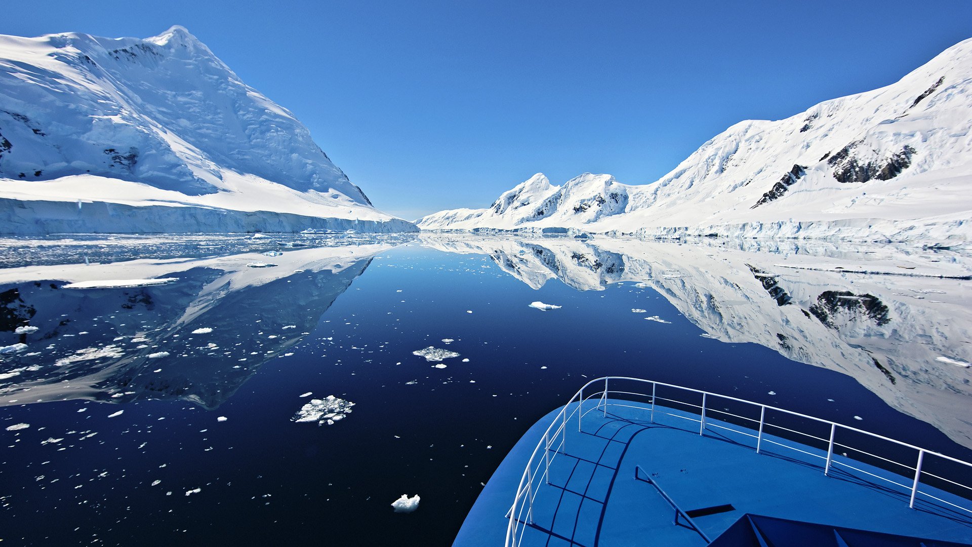 HD desktop wallpaper of a boat navigating calm Antarctic ocean waters, reflecting snowy mountains under a clear blue sky.