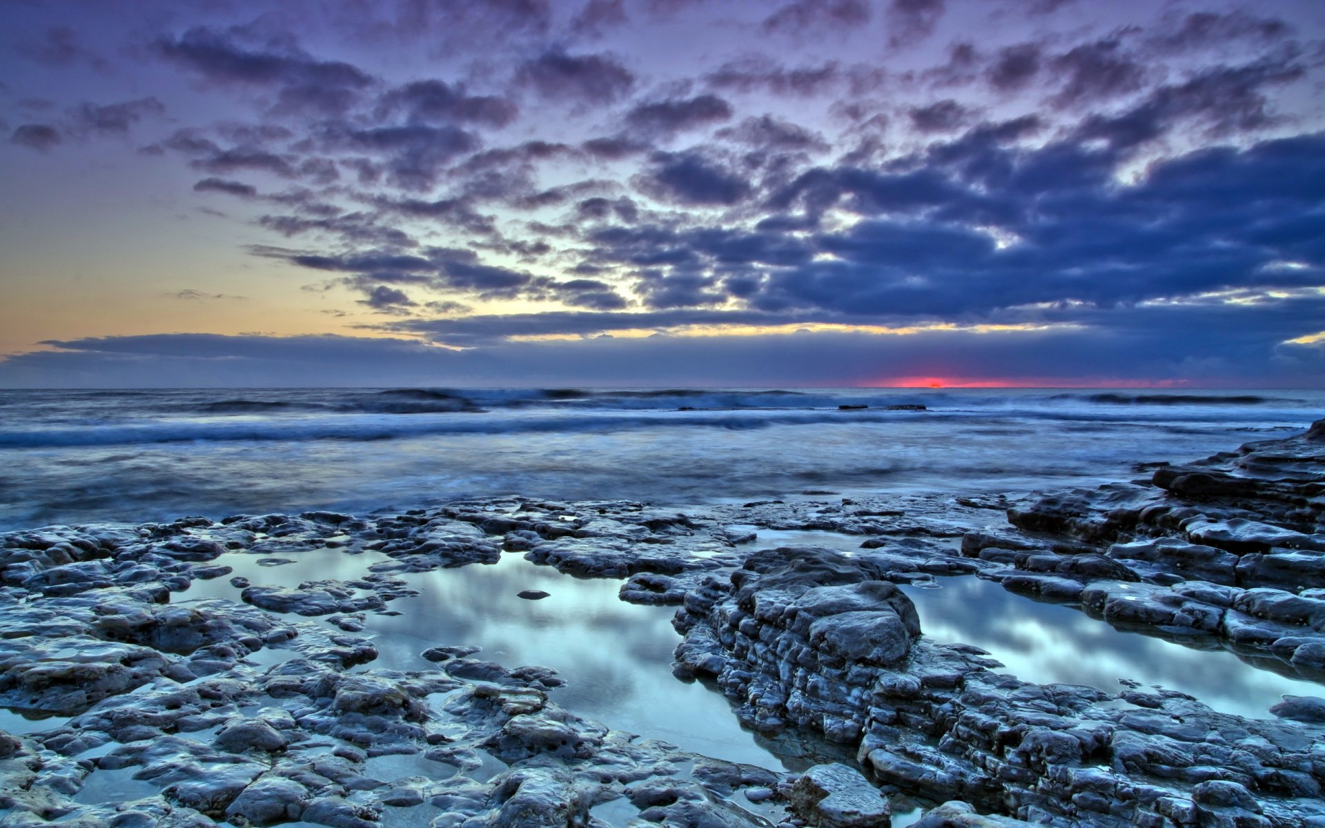 HD PC desktop wallpaper featuring a dramatic ocean scene with rocky shores under a vibrant, cloud-filled sky at sunset.