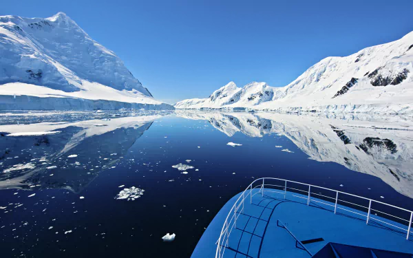 HD desktop wallpaper of a boat navigating calm Antarctic ocean waters, reflecting snowy mountains under a clear blue sky.