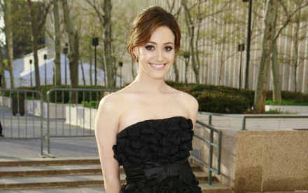 Emmy Rossum smiles elegantly in a stylish black dress, standing outdoors against a backdrop of trees, showcasing her poised demeanor in this HD desktop wallpaper.