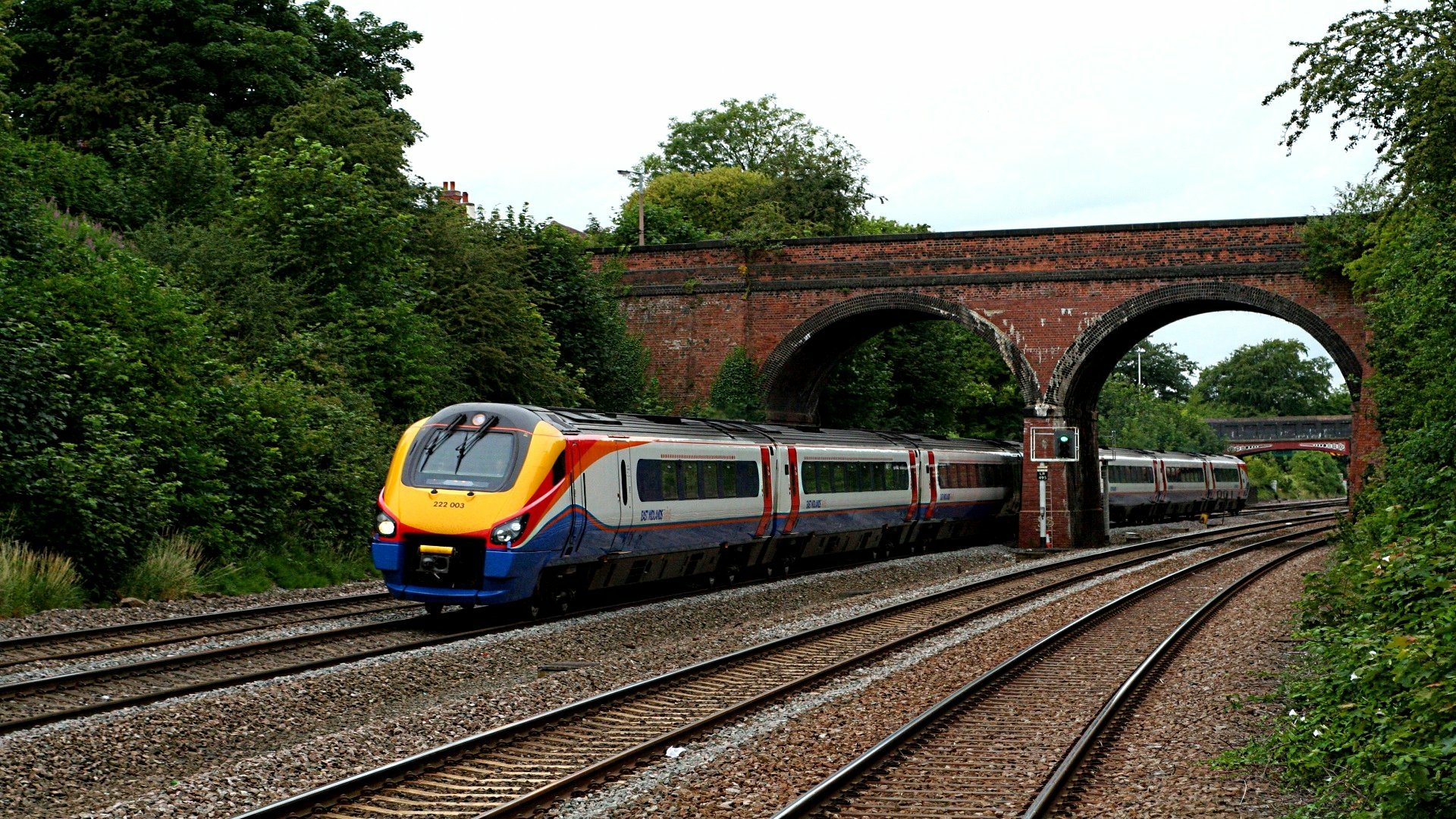 HD desktop wallpaper of a modern train traveling on tracks beneath a brick arch bridge, surrounded by lush green trees.