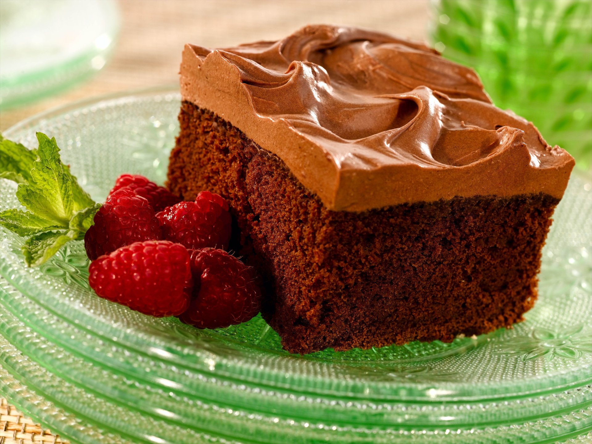 A rich chocolate cake slice topped with smooth frosting, served on a glass plate alongside fresh raspberries and a mint leaf, captured in vibrant 4K Ultra HD detail.