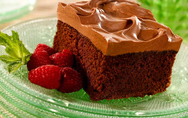 A rich chocolate cake slice topped with smooth frosting, served on a glass plate alongside fresh raspberries and a mint leaf, captured in vibrant 4K Ultra HD detail.