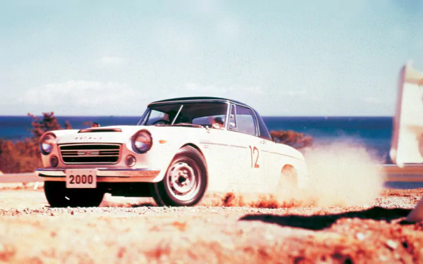 HD PC desktop wallpaper featuring a classic white Datsun vehicle speeding on a dusty road with the ocean and sky in the background.