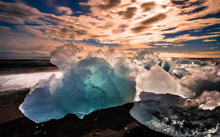 HD PC Desktop Wallpaper and Background — nature scene of translucent blue ice chunks on black sand, lit by a dramatic sunset and sweeping cloudscape.