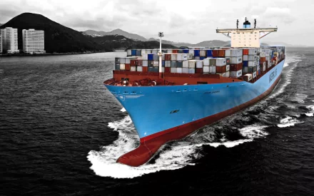 HD desktop wallpaper featuring a large container tanker vessel navigating through the water near a coastal city with mountainous background.