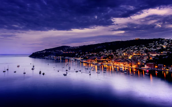 Stunning 4K Ultra HD view of Villefranche-sur-Mer, France, showcasing a serene harbor with boats and illuminated buildings under a dramatic sky.