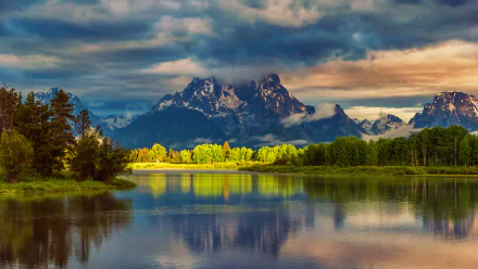 A serene river reflects majestic mountains under a moody sky, surrounded by lush greenery. This HD desktop wallpaper captures the beauty of nature perfectly.
