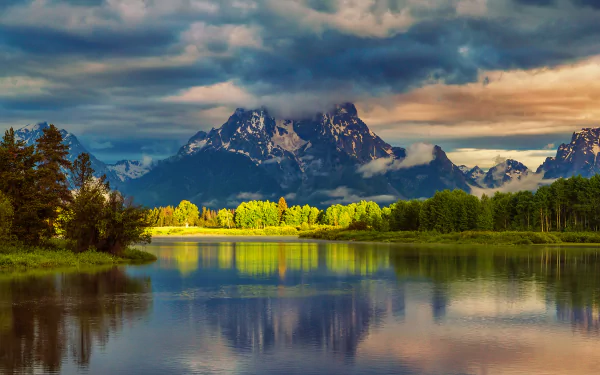 A serene river reflects majestic mountains under a moody sky, surrounded by lush greenery. This HD desktop wallpaper captures the beauty of nature perfectly.