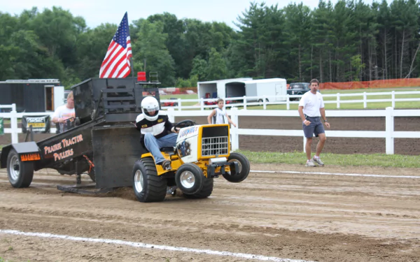 A vibrant HD wallpaper featuring a tractor pulling a load in a competition, with spectators watching and an American flag displayed, set against a green, wooded backdrop.