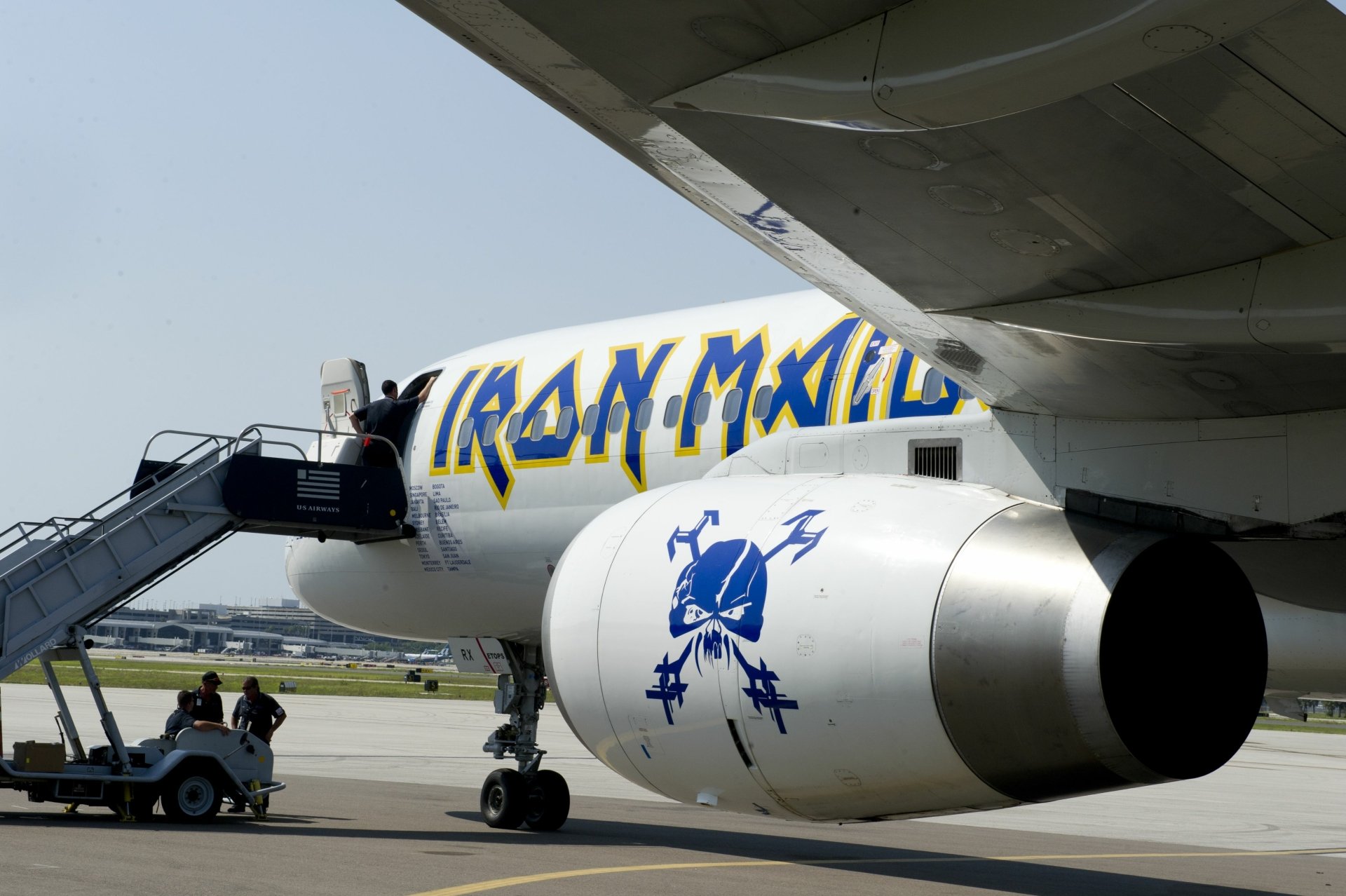 A vibrant 4K Ultra HD wallpaper featuring an Iron Maiden-branded airplane, showcasing the distinctive logo and artwork as crew members prepare for boarding on the tarmac.