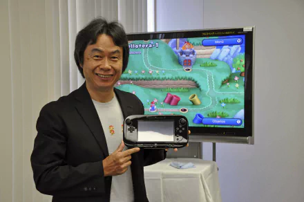 Man smiling and holding a gaming console with a game displayed on the screen, standing in front of a monitor, suitable as an HD desktop wallpaper and background.