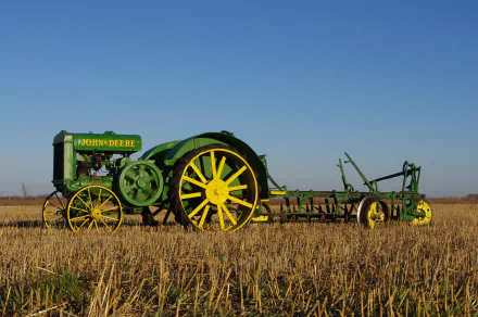 A vintage John Deere tractor stands prominently in a golden field, showcasing its distinct green and yellow colors against a clear blue sky. 4K Ultra HD desktop wallpaper and background.
