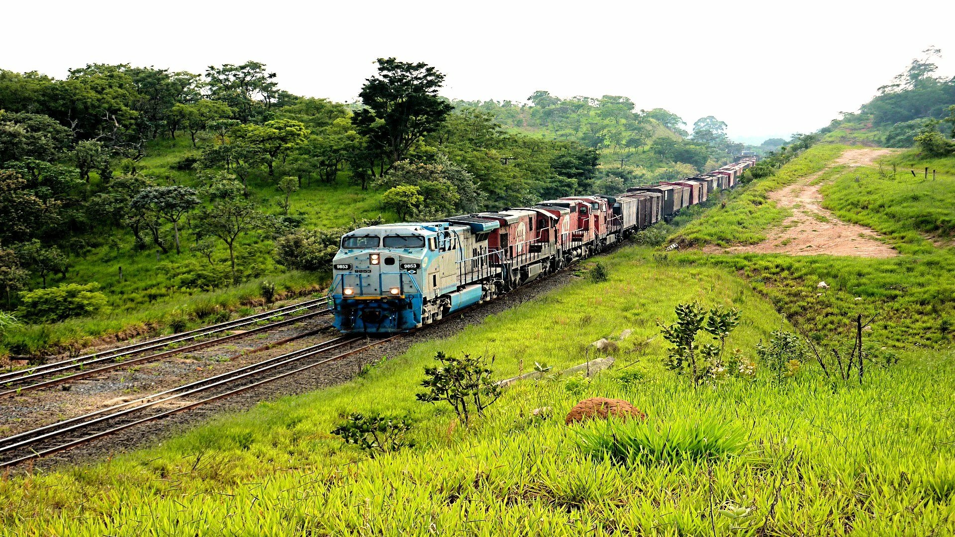 HD desktop wallpaper of a freight train traveling through lush green countryside on parallel tracks under a bright sky.