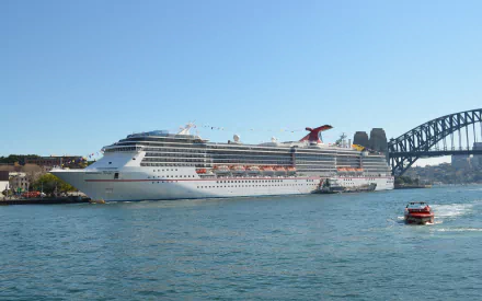 A panoramic view of Sydney Harbour featuring the Carnival Spirit cruise ship docked beside the iconic Sydney Harbour Bridge, with a smaller boat navigating the sparkling waters.