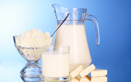 HD desktop wallpaper featuring a glass pitcher filled with milk, accompanied by a bowl of cottage cheese, a glass of milk, and several pieces of white cheese, set against a gradient blue background.