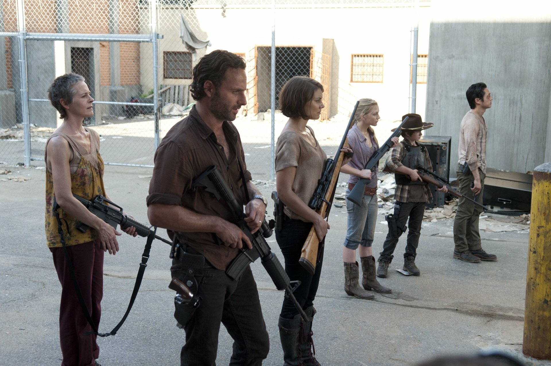 A group of survivors from The Walking Dead, including Rick Grimes, Maggie Greene, and Glenn Rhee, stands ready for a confrontation in a desolate urban setting.