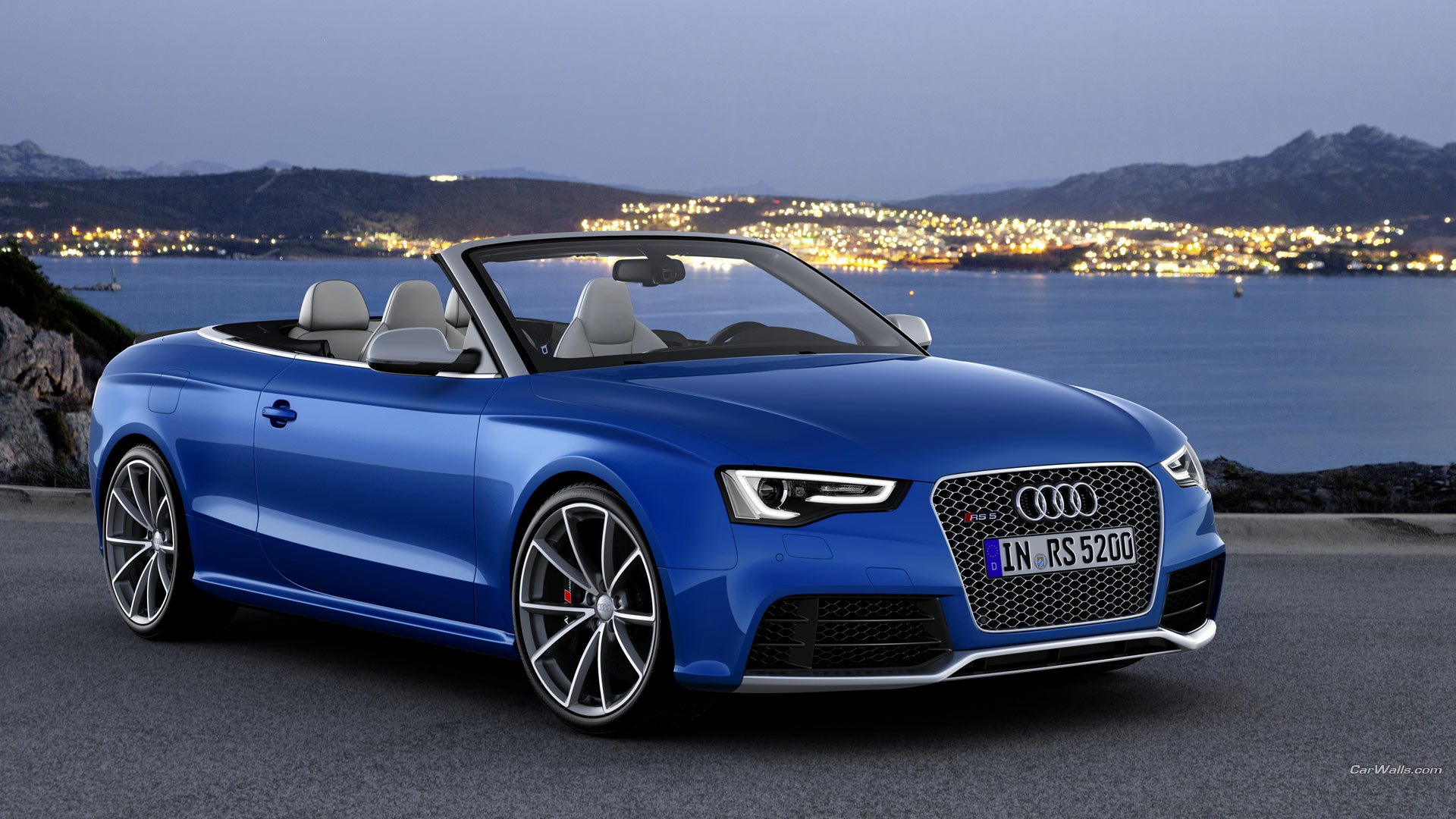 HD PC desktop wallpaper featuring a sleek blue Audi RS5 convertible parked near a body of water with city lights and hills in the background at dusk.