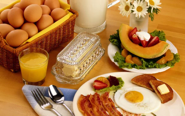 HD desktop wallpaper featuring a breakfast spread with eggs, bacon, toast with butter, juice, milk, fresh fruit, and a basket of eggs on a wooden table.