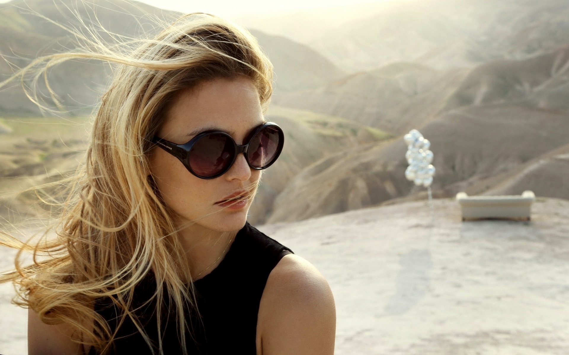 2K Quad HD desktop wallpaper: celebrity portrait of a woman in oversized sunglasses and a black sleeveless top, windblown hair, gazing over sunlit desert hills.