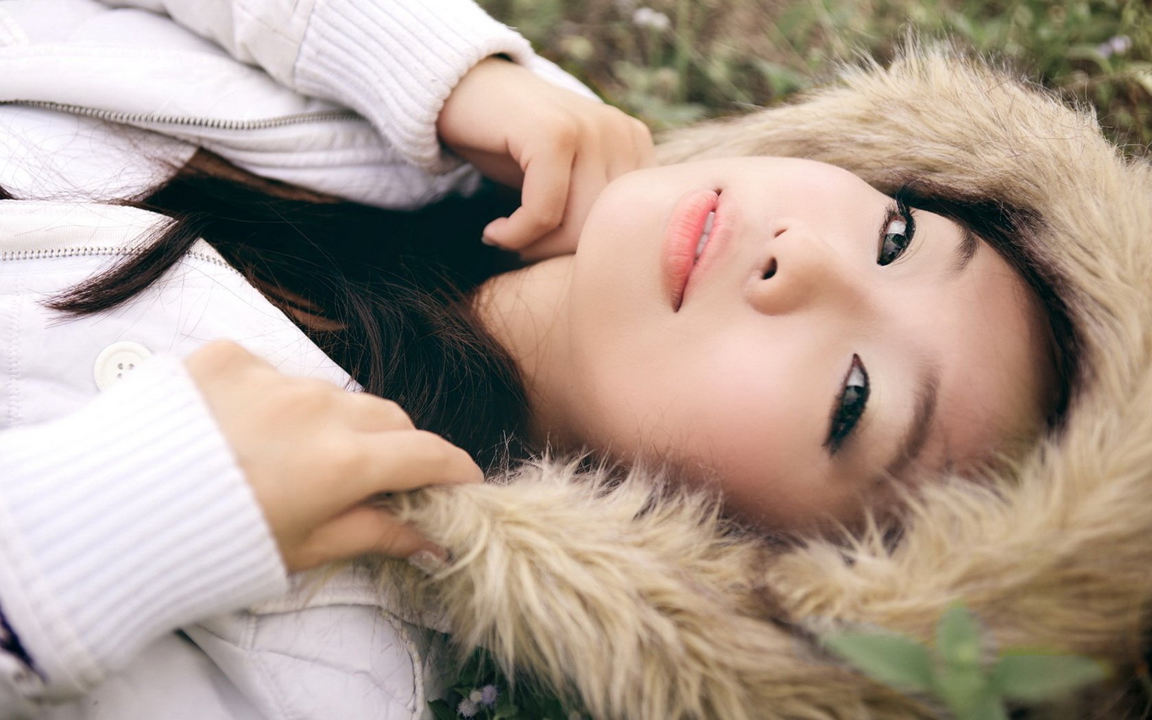 HD desktop wallpaper featuring a model laying on grass, wearing a white jacket with a fur-lined hood.