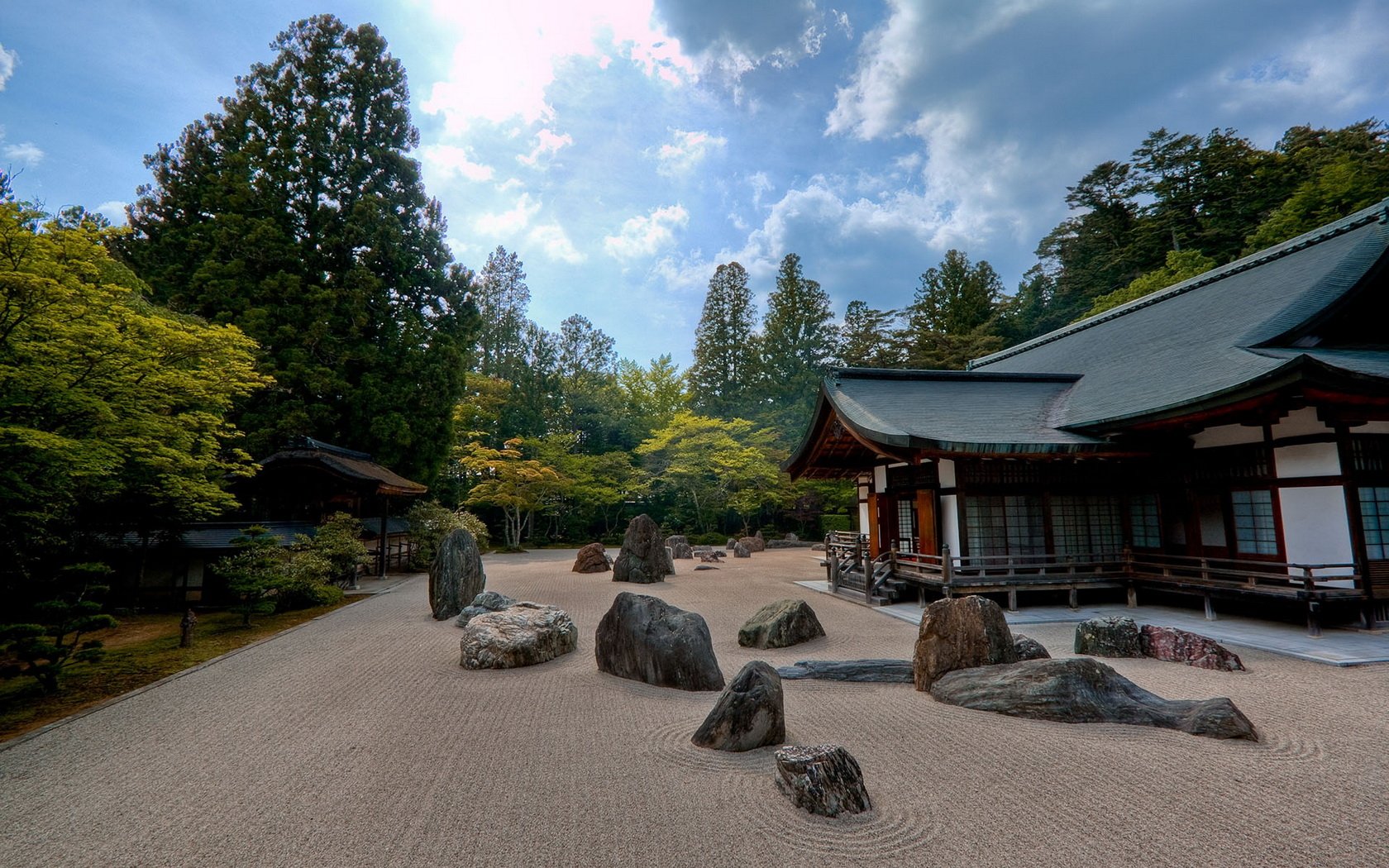 HD PC desktop wallpaper of a man-made rock garden with carefully arranged stones, surrounded by trees and traditional wooden architecture under a partly cloudy sky.