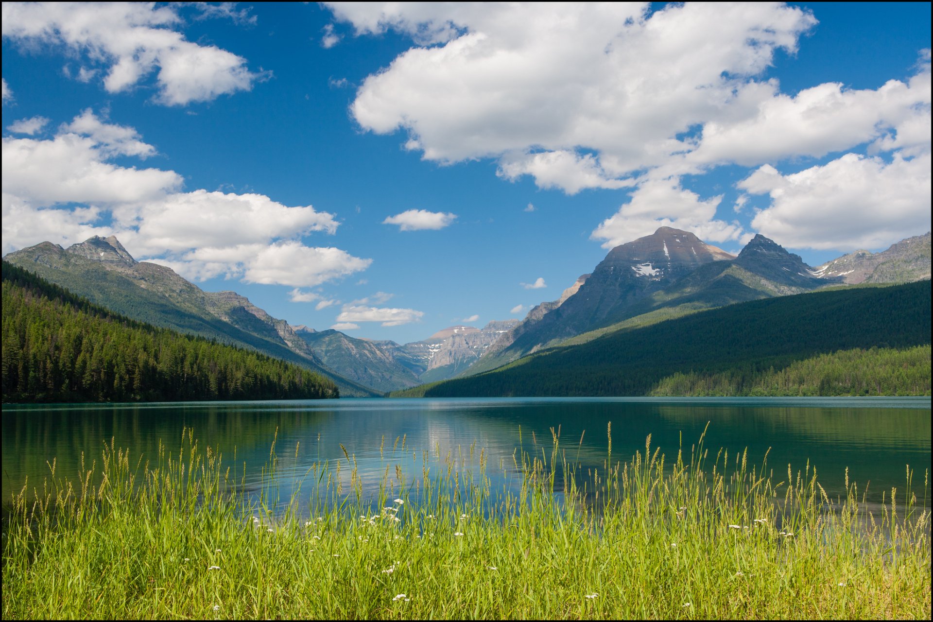 Scenic HD desktop wallpaper of Lake McDonald featuring a tranquil lake, lush green landscape, blue sky with clouds, and distant mountains under the sun.