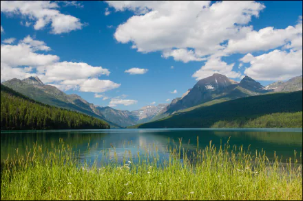 Scenic HD desktop wallpaper of Lake McDonald featuring a tranquil lake, lush green landscape, blue sky with clouds, and distant mountains under the sun.