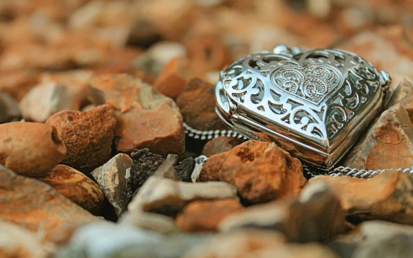 A silver heart-shaped locket rests among pebbles, showcasing intricate designs. This HD wallpaper captures the beauty of man-made jewelry in a natural setting.